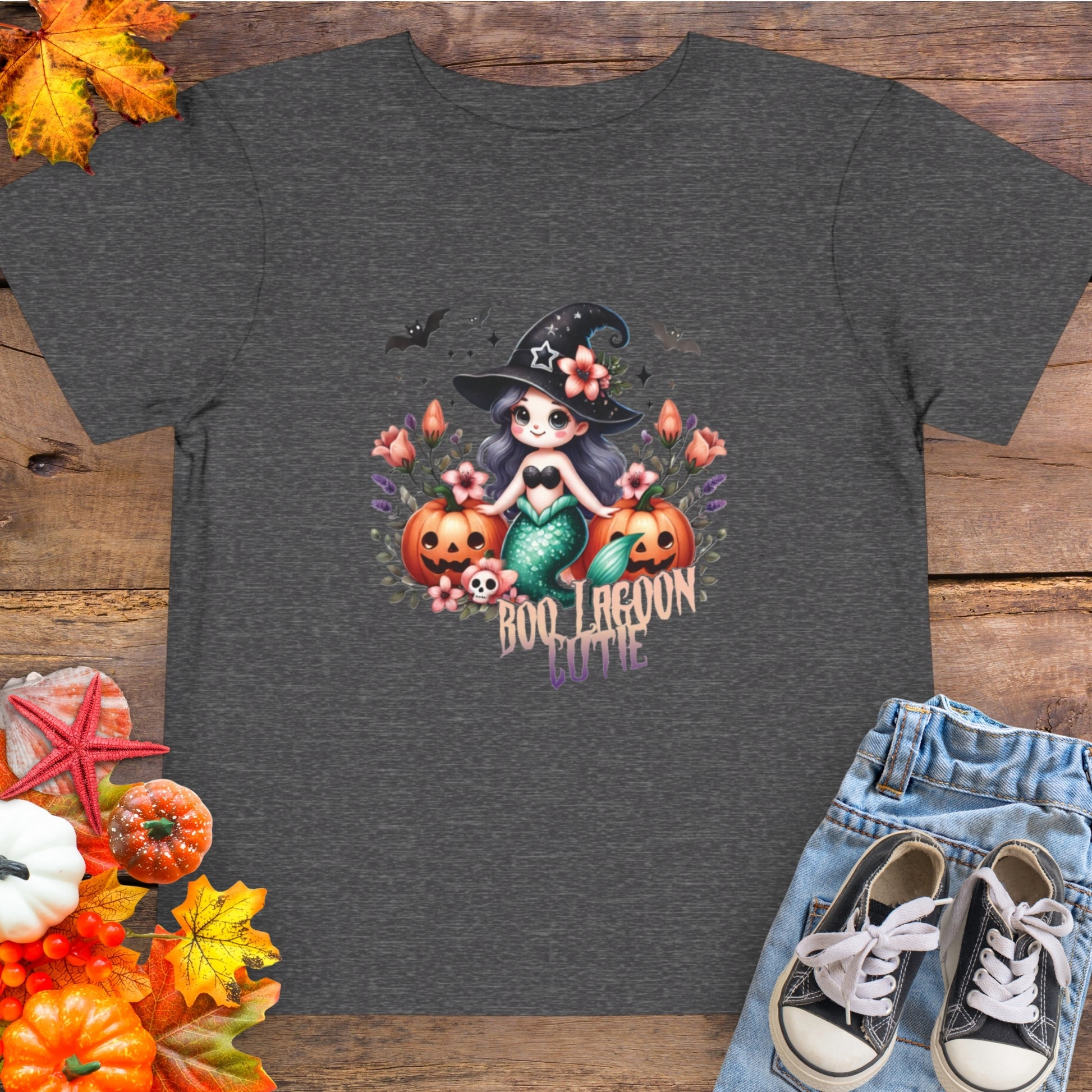 Dark gray Toddler t-shirt design with a witchy mermaid sitting among pumpkins and flowers, with bats flying overhead and the words “Boo Lagoon Cutie.