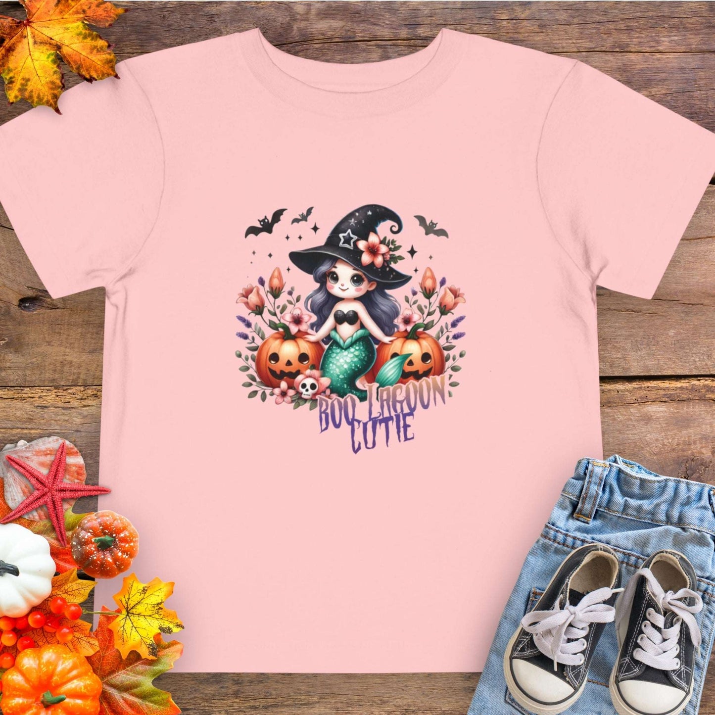 Pink Toddler t-shirt design with a witchy mermaid sitting among pumpkins and flowers, with bats flying overhead and the words “Boo Lagoon Cutie.