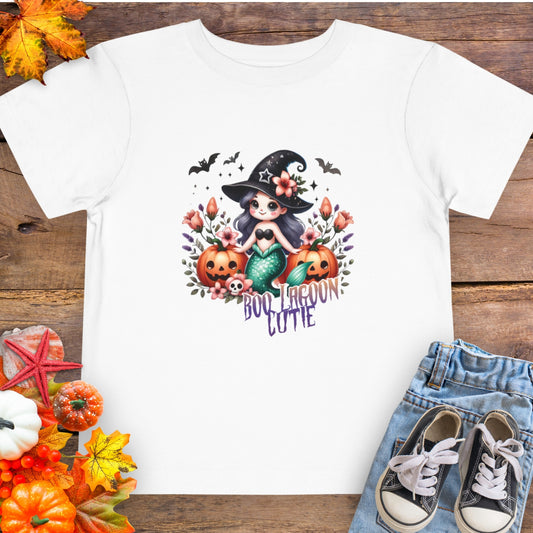 White Toddler t-shirt design with a witchy mermaid sitting among pumpkins and flowers, with bats flying overhead and the words “Boo Lagoon Cutie.