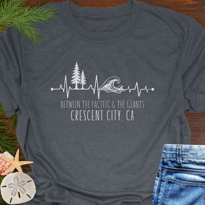 Gray t-shirt with a design of trees and  wave on a heartline, and text 'Between the Pacific and Giants Crescent City, CA'.