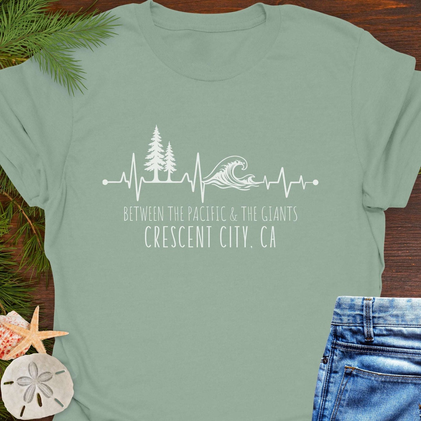 Green t-shirt with a design of trees and  wave on a heartline, and text 'Between the Pacific and Giants Crescent City, CA'.