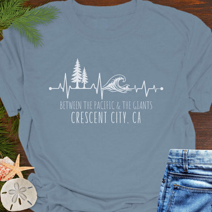 Blue t-shirt with a design of trees and  wave on a heartline, and text 'Between the Pacific and Giants Crescent City, CA'.