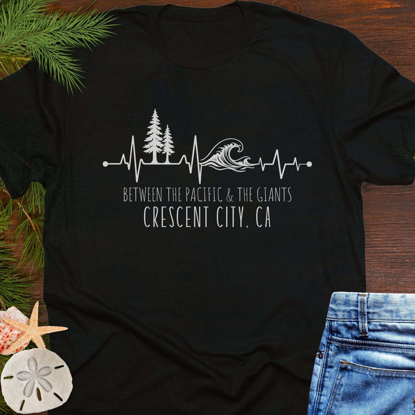 Black t-shirt with a design of trees and  wave on a heartline, and text 'Between the Pacific and Giants Crescent City, CA'.
