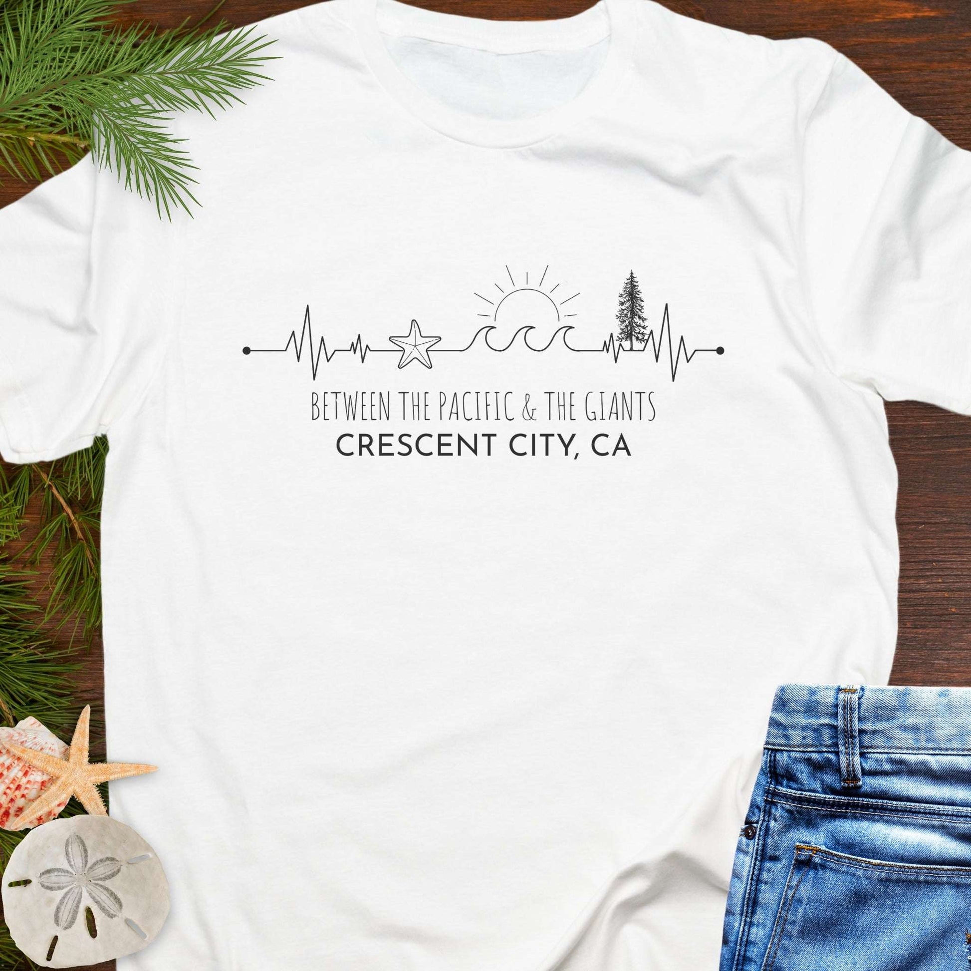 White t-shirt with a design of trees and  wave on a heartline, and text 'Between the Pacific and Giants Crescent City, CA'.