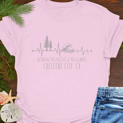 Pink t-shirt with a design of trees and  wave on a heartline, and text 'Between the Pacific and Giants Crescent City, CA'.