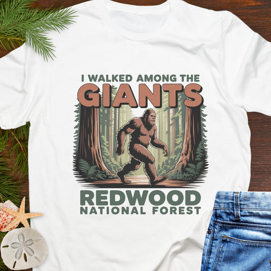 White t-shirt with a Sasquatch graphic and text 'I Walked Among the Giants Redwood National Forest' on a wooden surface.