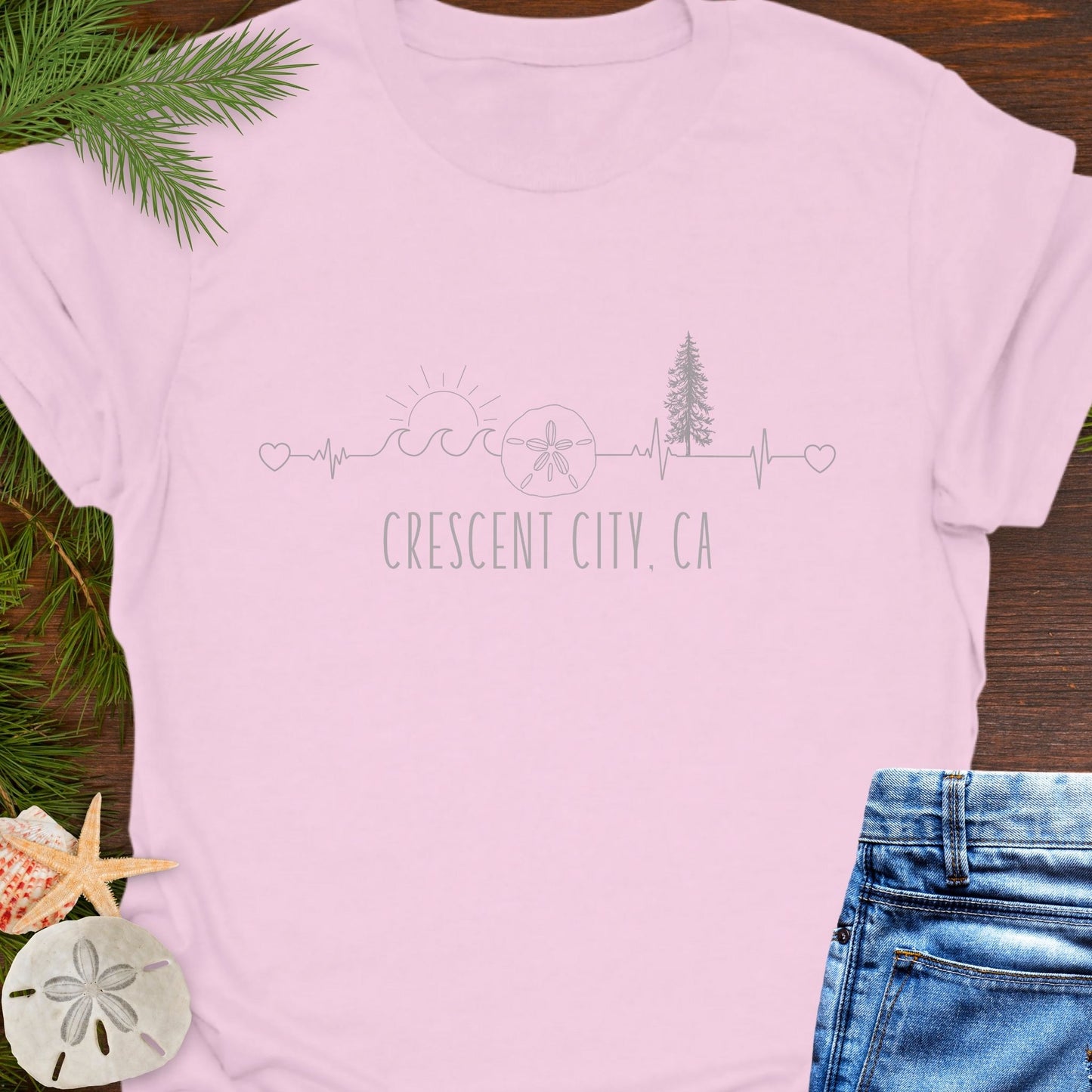 Pink t-shirt with white heartline with redwoods, waves, and sand dollar with text 'Crescent City, CA'.