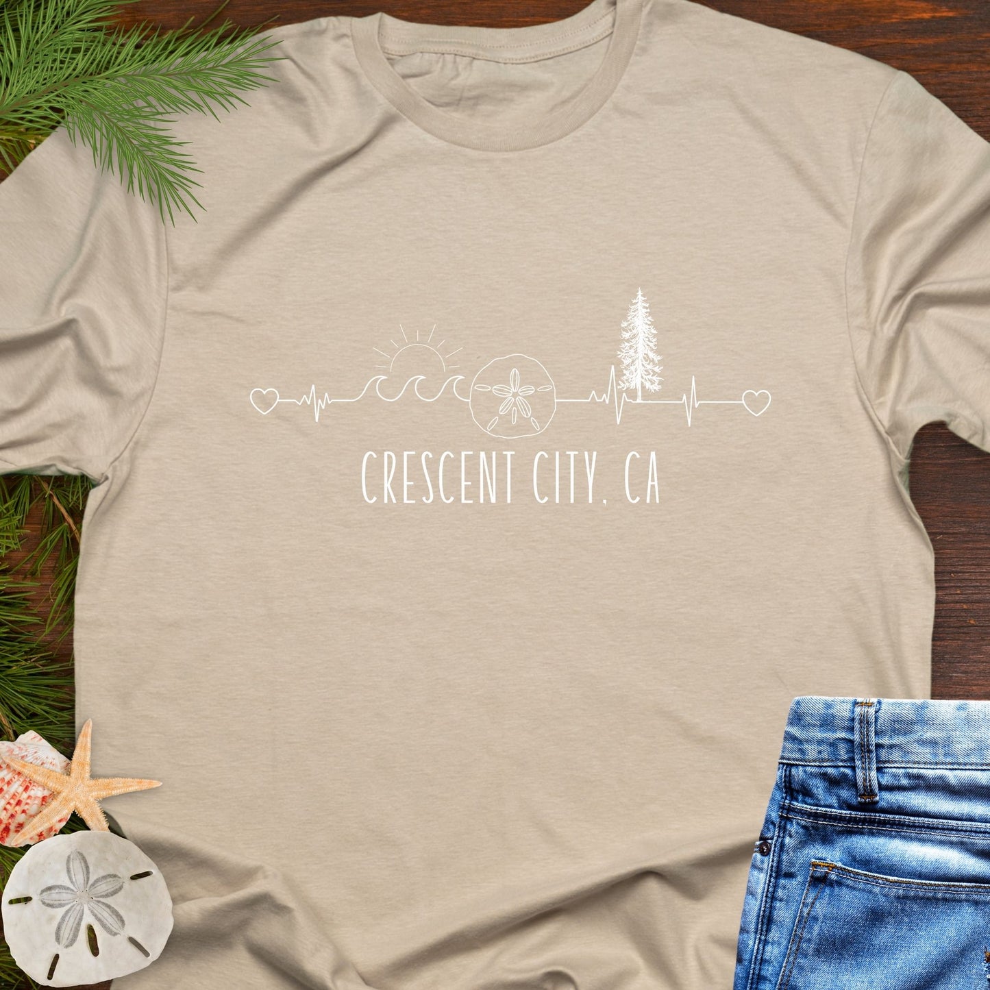 Beige t-shirt with white heartline with redwoods, waves, and sand dollar with text 'Crescent City, CA'.