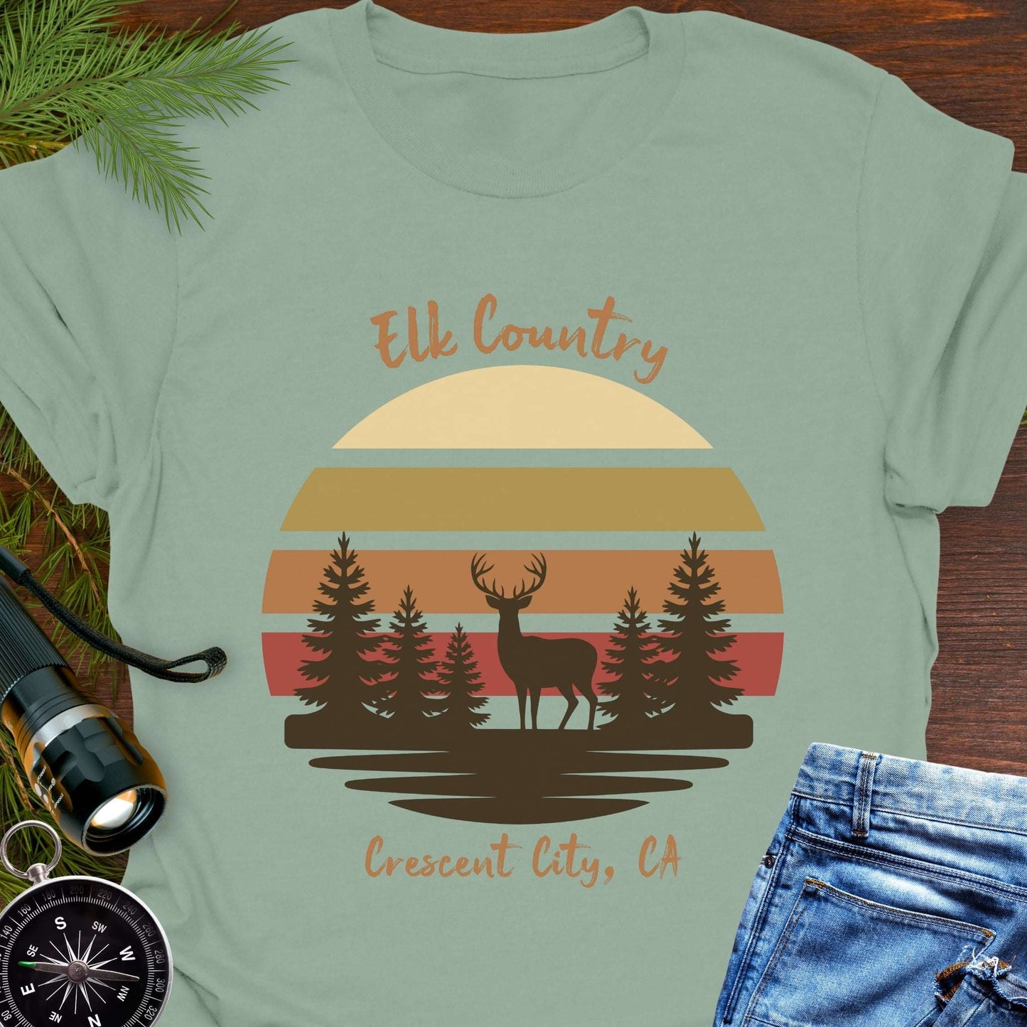 Light green t-shirt with muted retro sunset with brown silhouette deer and trees with text 'Elk country Crescent City, CA.
