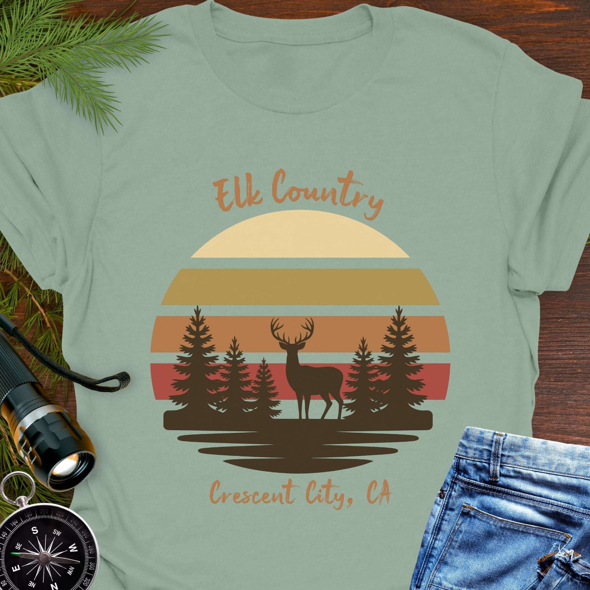 Light green t-shirt with muted retro sunset with brown silhouette deer and trees with text 'Elk country Crescent City, CA.