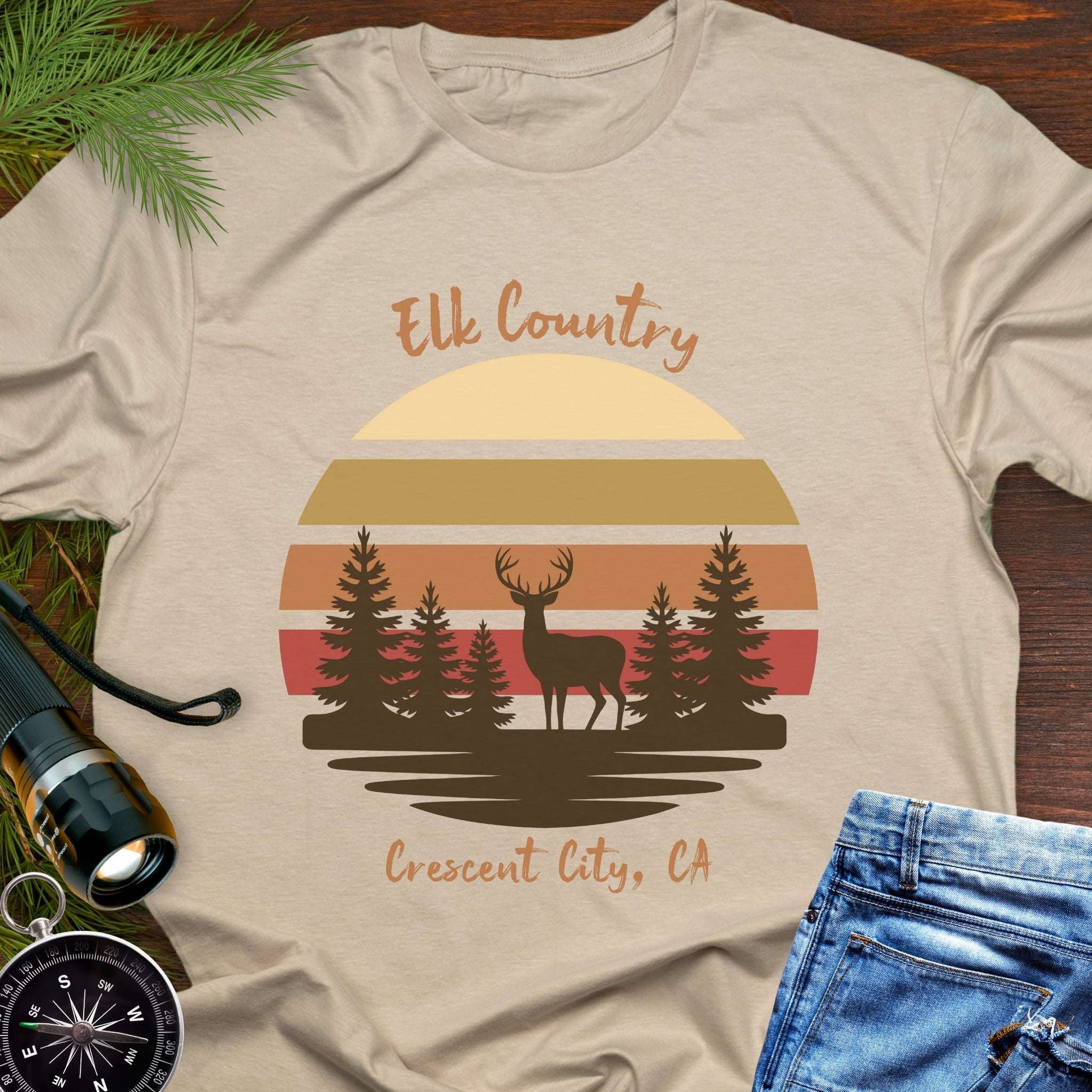 Beige t-shirt with muted retro sunset with brown silhouette deer and trees with text 'Elk country Crescent City, CA.