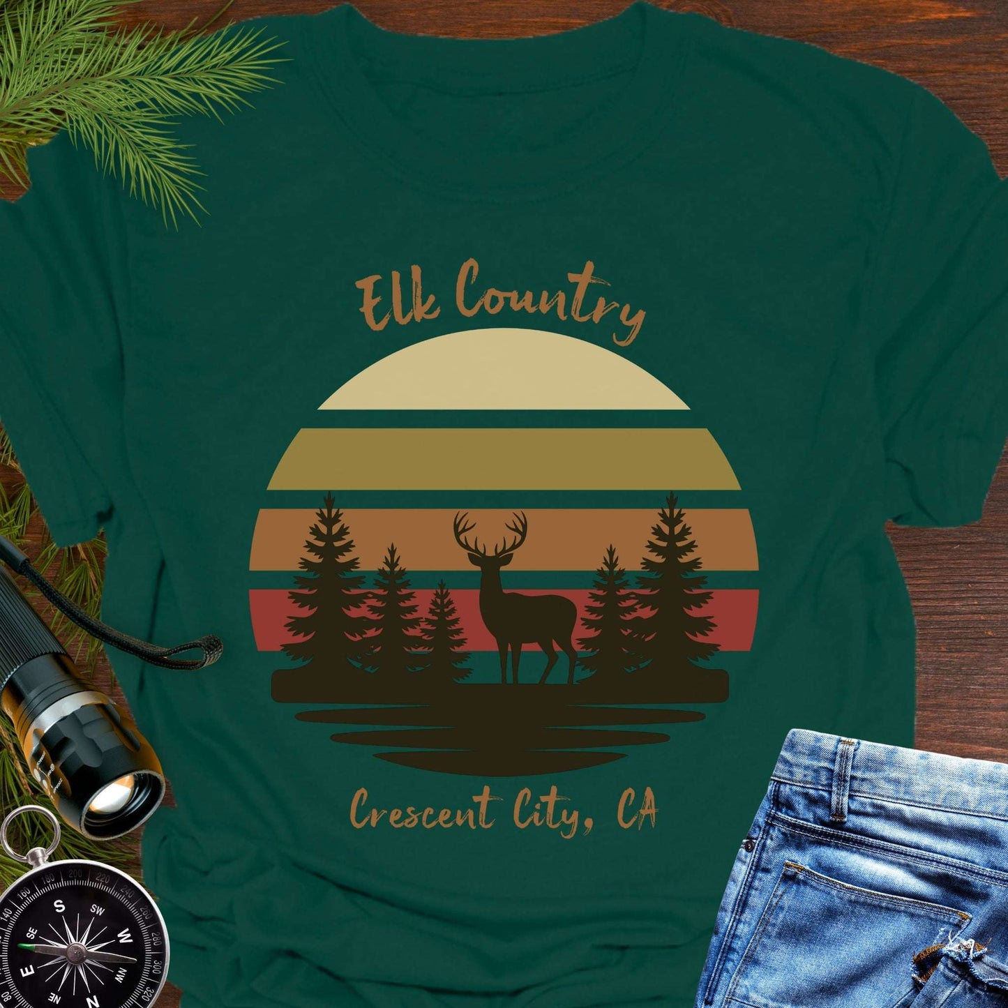 Green t-shirt with muted retro sunset with brown silhouette deer and trees with text 'Elk country Crescent City, CA.