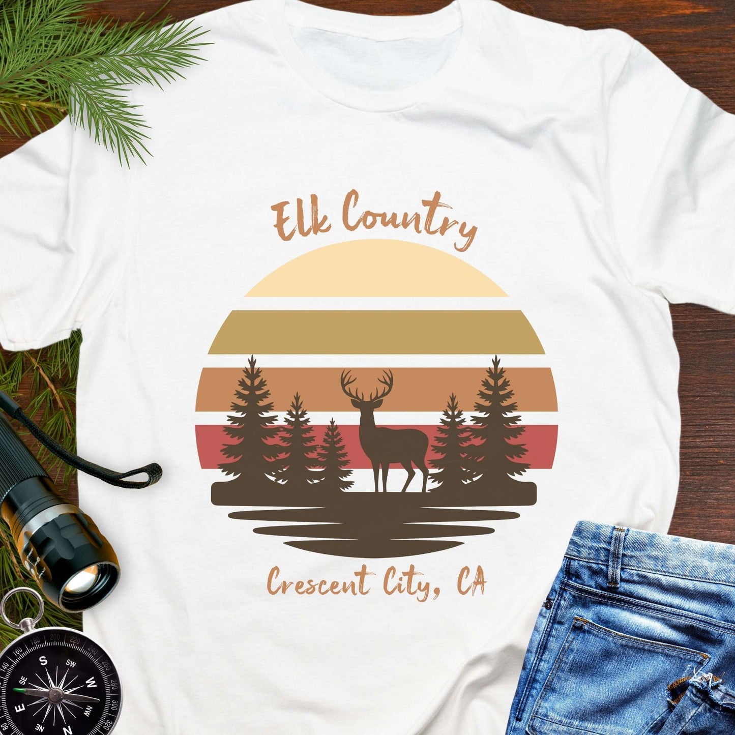 White t-shirt with muted retro sunset with brown silhouette deer and trees with text 'Elk country Crescent City, CA.