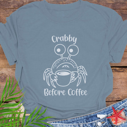 Blue t-shirt with a crab graphic and text 'Crabby Before Coffee' on a wooden surface.