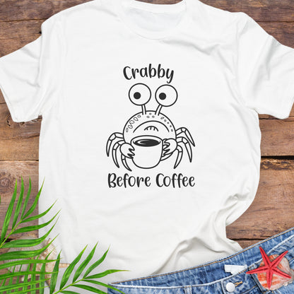 White t-shirt with a crab graphic and 'Crabby Before Coffee' text on a wooden surface with leaves and jeans.