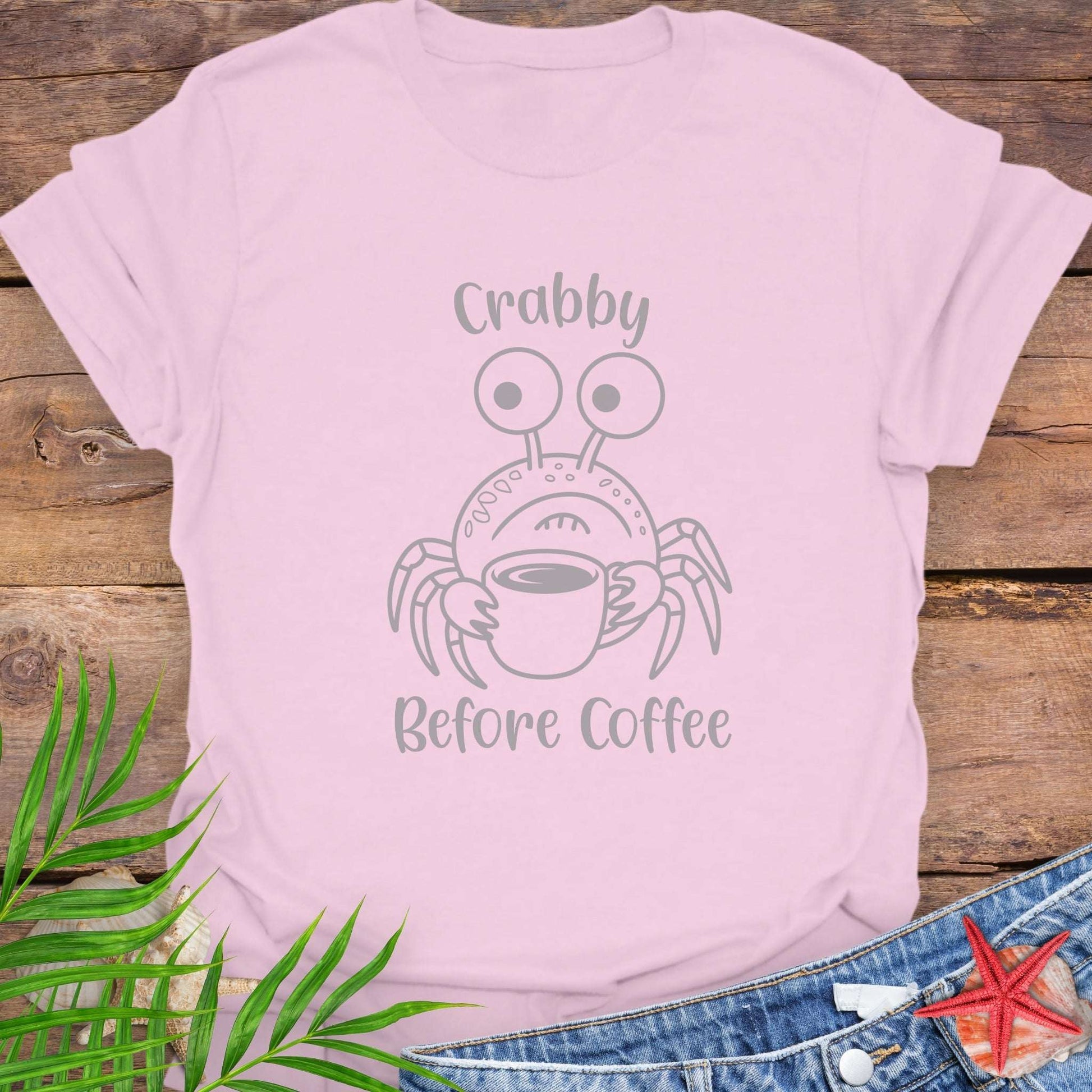 Pink t-shirt with crab graphic and text 'Crabby Before Coffee' on a wooden surface with leaves and jeans.