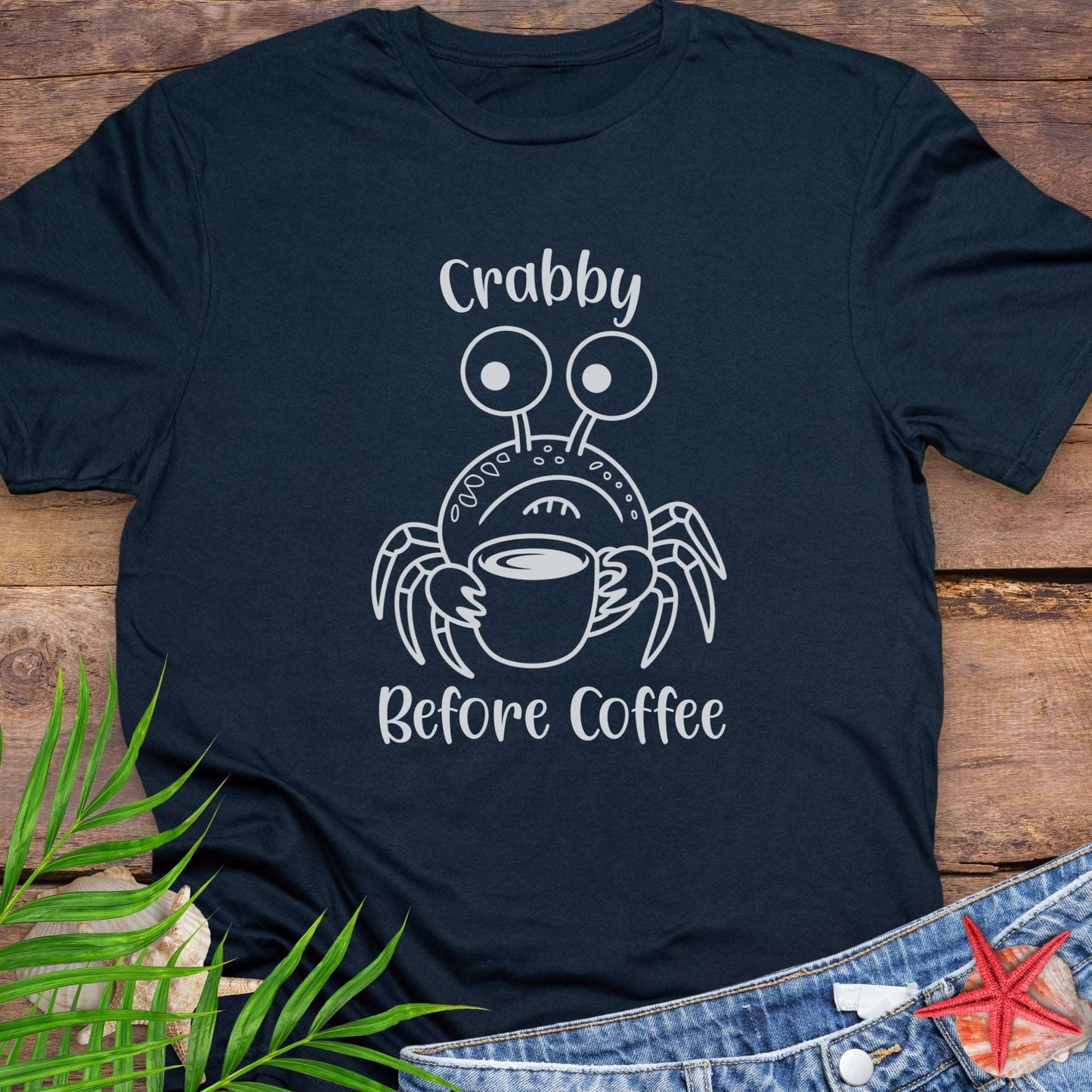 Navy blue t-shirt with a crab graphic and text 'Crabby Before Coffee' on a wooden surface.
