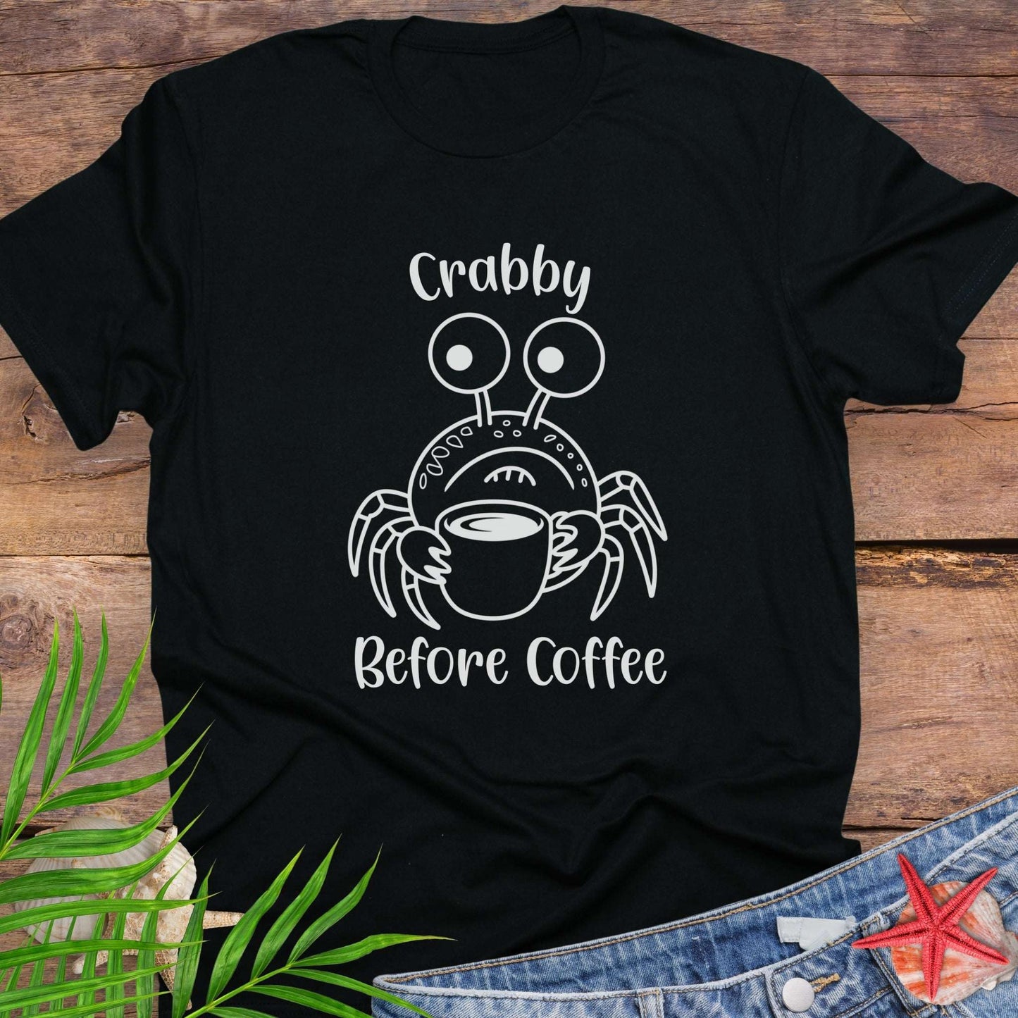 Black t-shirt with a crab graphic and text 'Crabby Before Coffee' on a wooden surface.