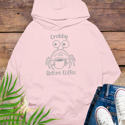 Pink hoodie with crab graphic and text 'Crabby Before Coffee' on a wooden surface with jeans and sneakers.