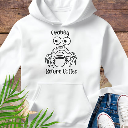 White hoodie with a crab graphic and text 'Crabby Before Coffee' on a wooden surface.