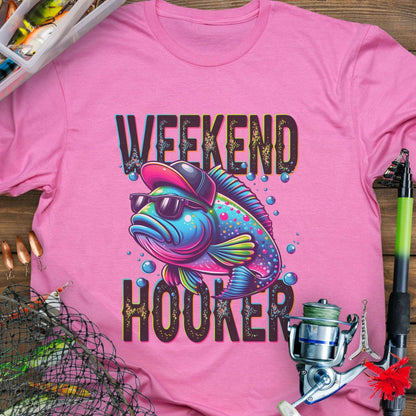 Pink t-shirt with a colorful fish graphic and 'Weekend Hooker' text, surrounded by fishing equipment on a wooden surface.