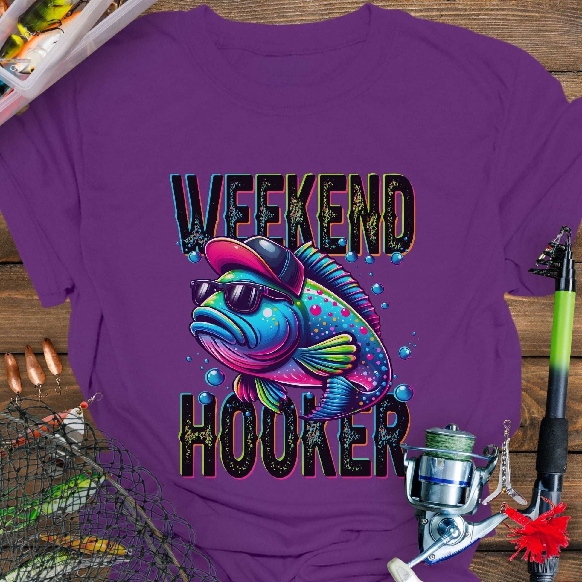 Purple t-shirt with a colorful fish graphic and 'Weekend Hooker' text, surrounded by fishing equipment on a wooden surface.