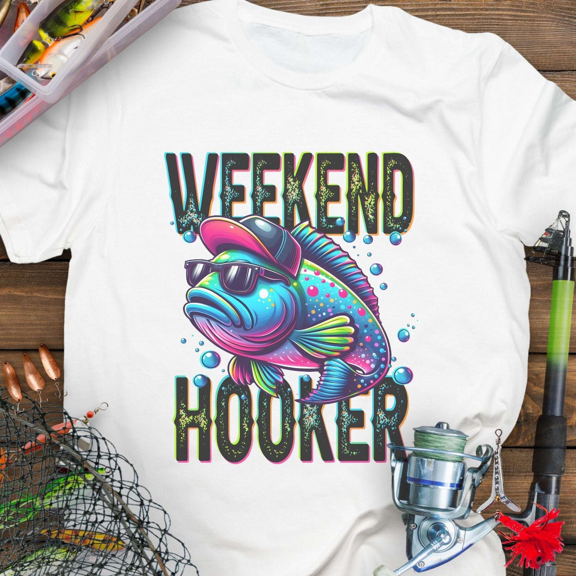 White t-shirt with colorful fish graphic and 'Weekend Hooker' text on a wooden surface with fishing equipment.