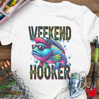 White t-shirt with colorful fish graphic and 'Weekend Hooker' text on a wooden surface with fishing equipment.
