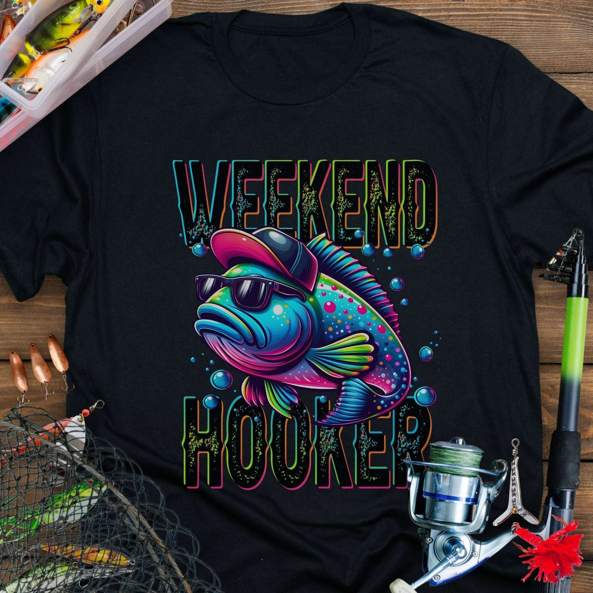 Black t-shirt with a colorful fish graphic and 'Weekend Hooked' text on a wooden surface with fishing equipment.