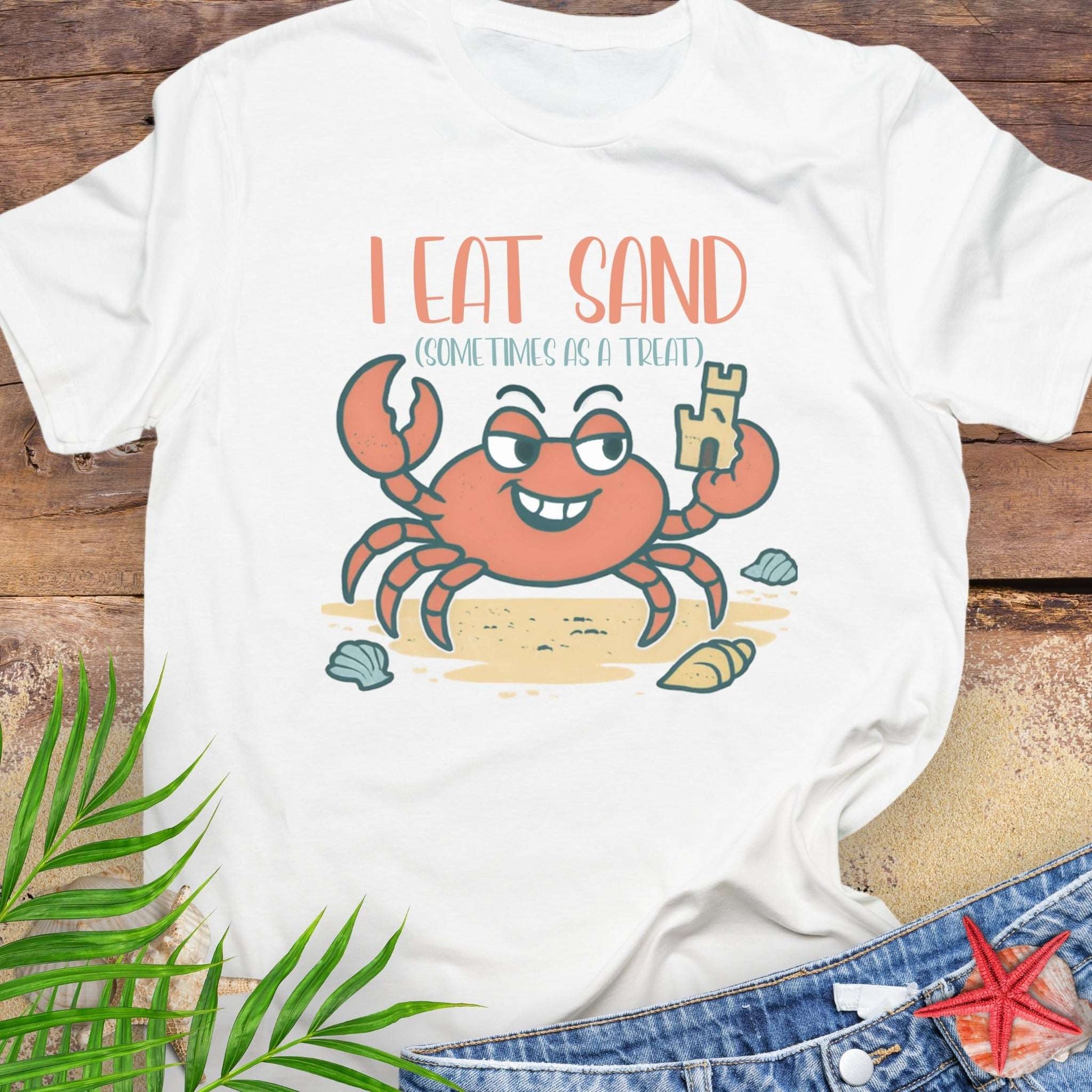 White t-shirt with a cartoon crab design and text 'I Eat Sand Sometimes as a Treat' on a wooden surface.