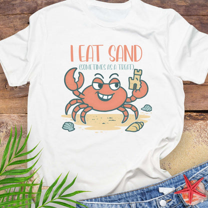 White t-shirt with a cartoon crab design and text 'I Eat Sand Sometimes as a Treat' on a wooden surface.