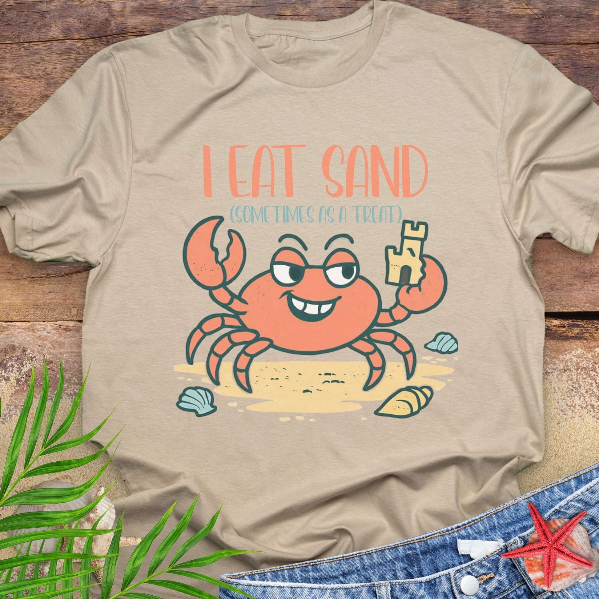 Beige t-shirt with a cartoon crab and 'I Eat Sand' text on a wooden surface.