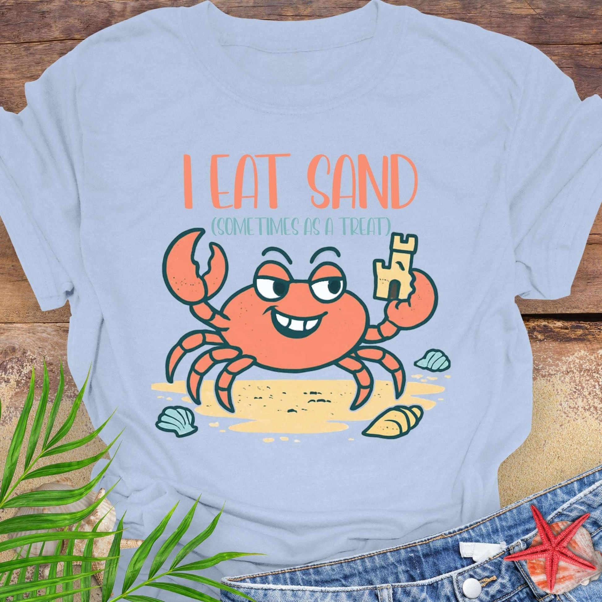 Light blue t-shirt with a cartoon crab design and text 'I Eat Sand Sometimes as a Treat' on a wooden surface.