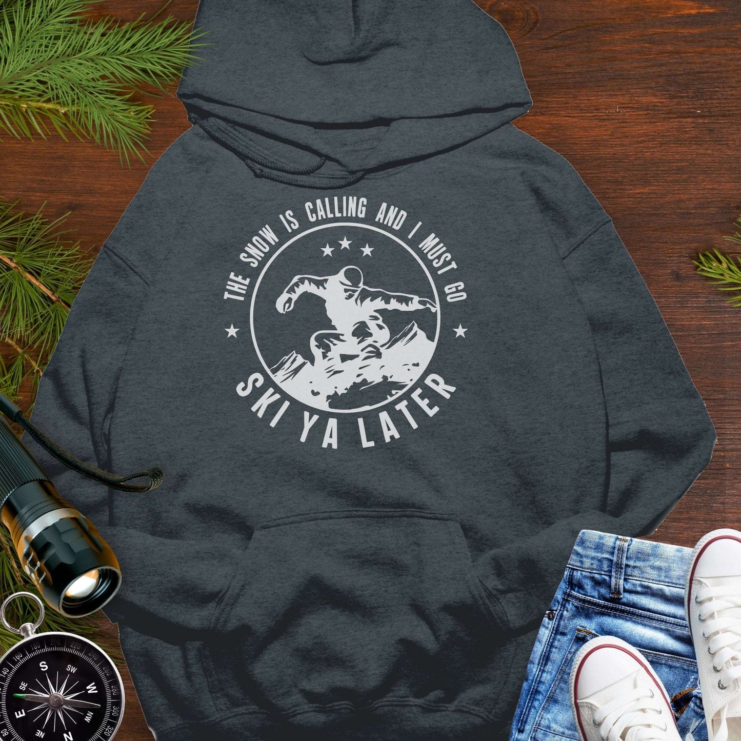 Dark gray hoodie with 'Ski Ya Later' graphic on a wooden surface with winter accessories.