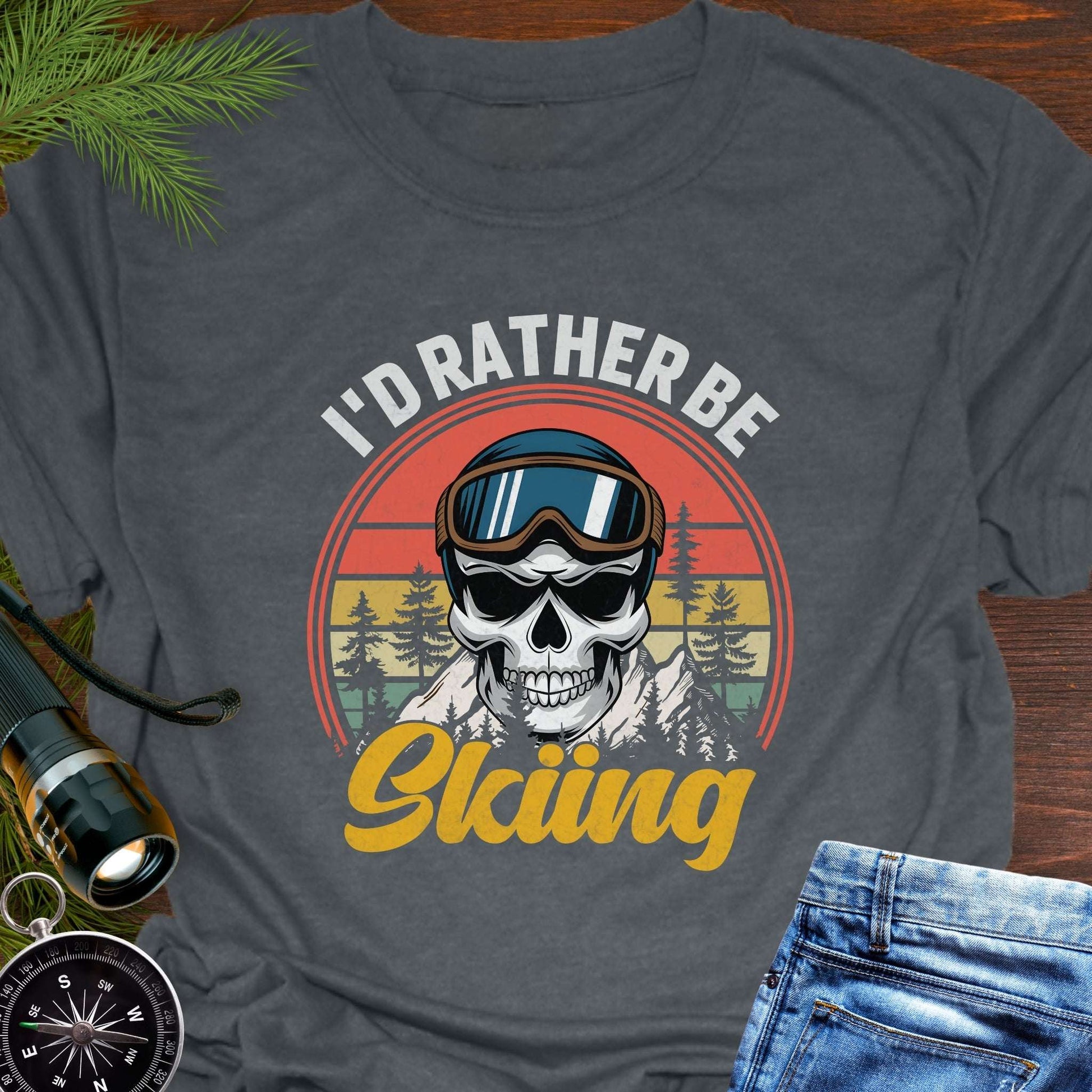 Gray T-shirt graphic showing a skeleton head wearing ski goggles in front of a retro sunset with mountains and trees, with text that reads “I’d Rather Be Skiing.