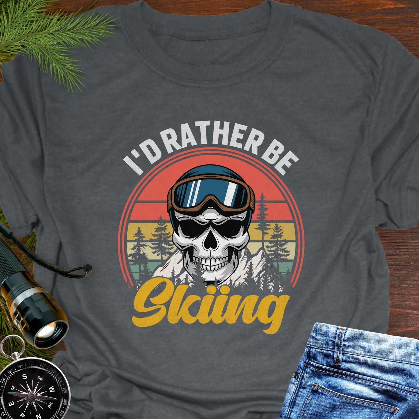 Gray T-shirt graphic showing a skeleton head wearing ski goggles in front of a retro sunset with mountains and trees, with text that reads “I’d Rather Be Skiing.