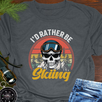 Gray T-shirt graphic showing a skeleton head wearing ski goggles in front of a retro sunset with mountains and trees, with text that reads “I’d Rather Be Skiing.