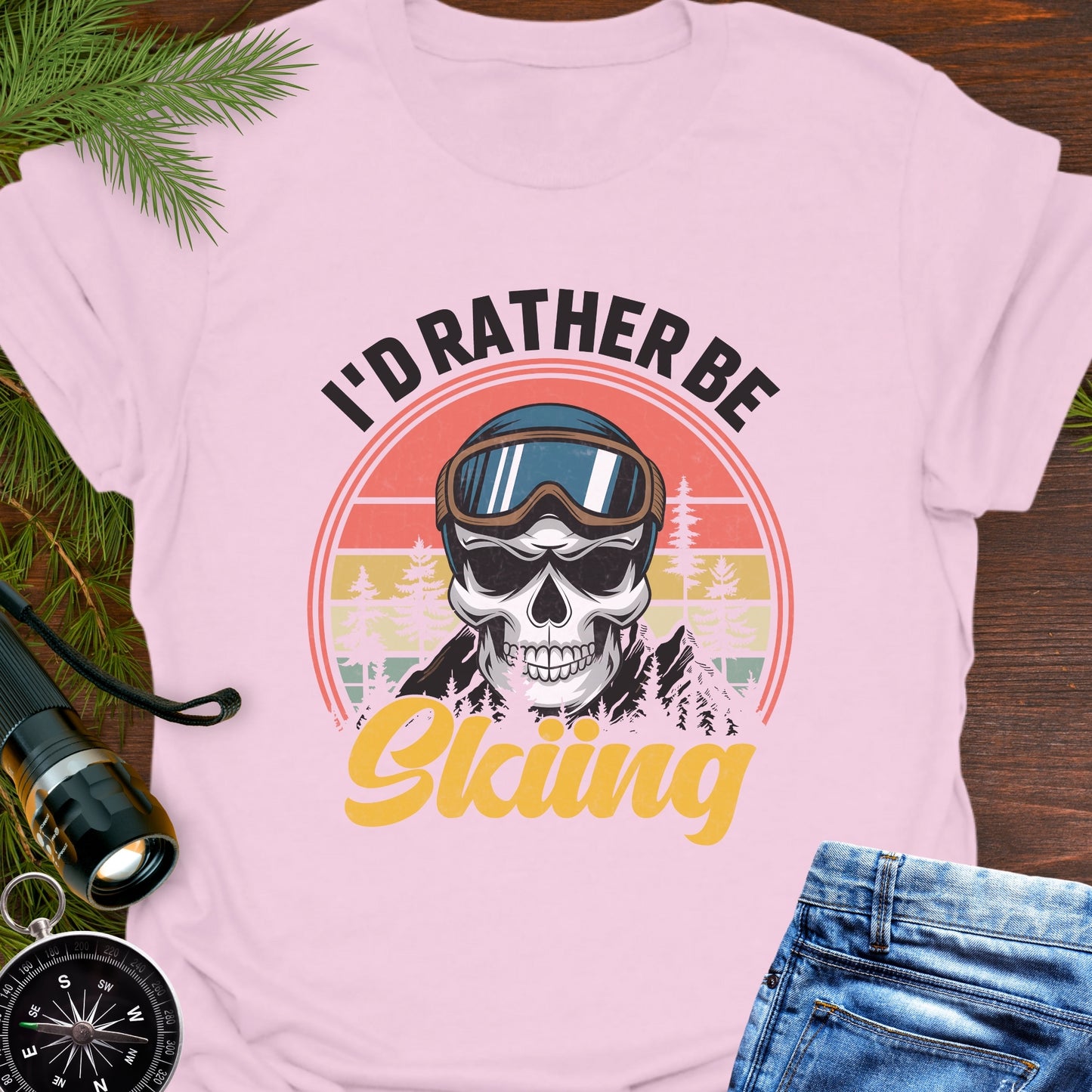 Pink T-shirt graphic showing a skeleton head wearing ski goggles in front of a retro sunset with mountains and trees, with text that reads “I’d Rather Be Skiing.