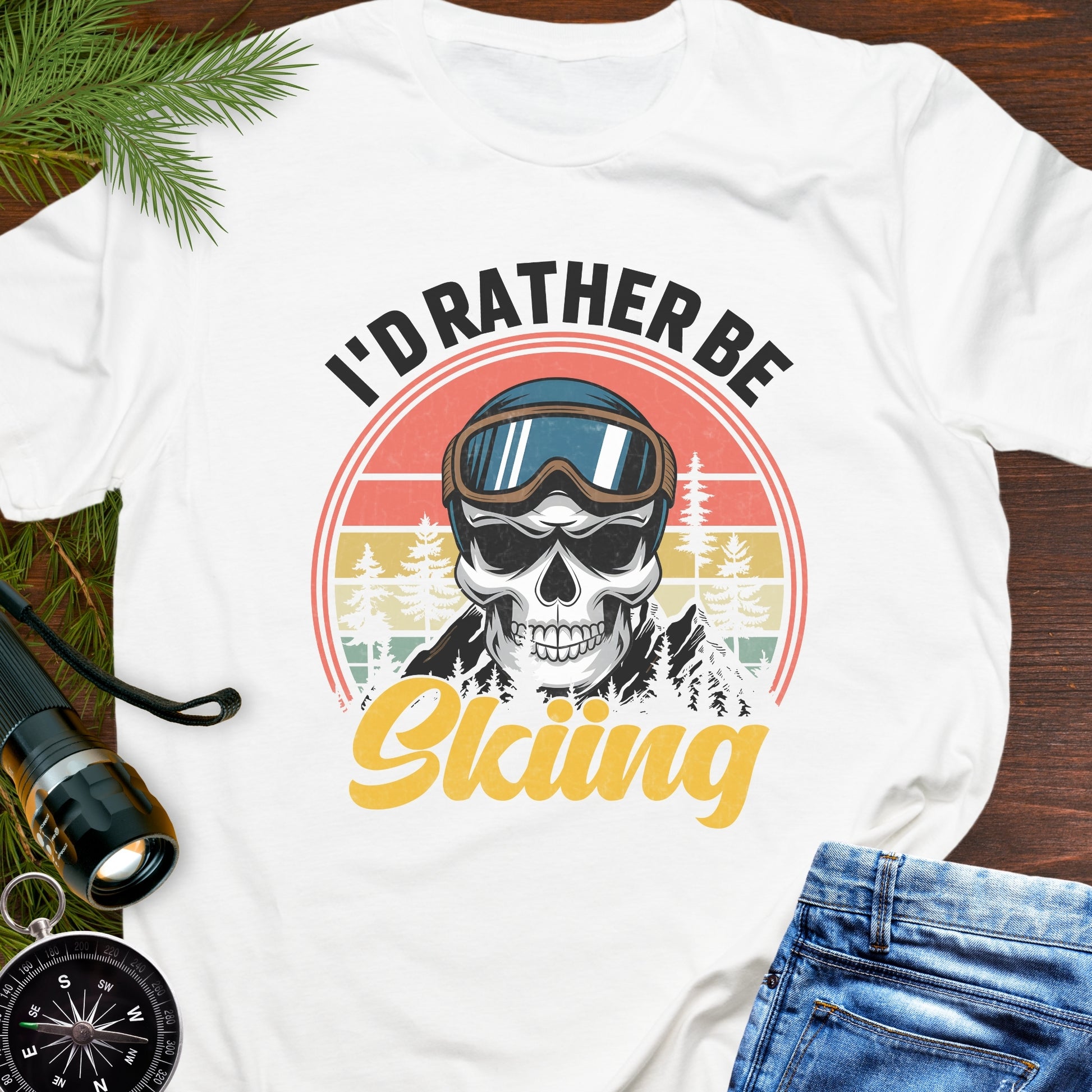 White T-shirt graphic showing a skeleton head wearing ski goggles in front of a retro sunset with mountains and trees, with text that reads “I’d Rather Be Skiing.
