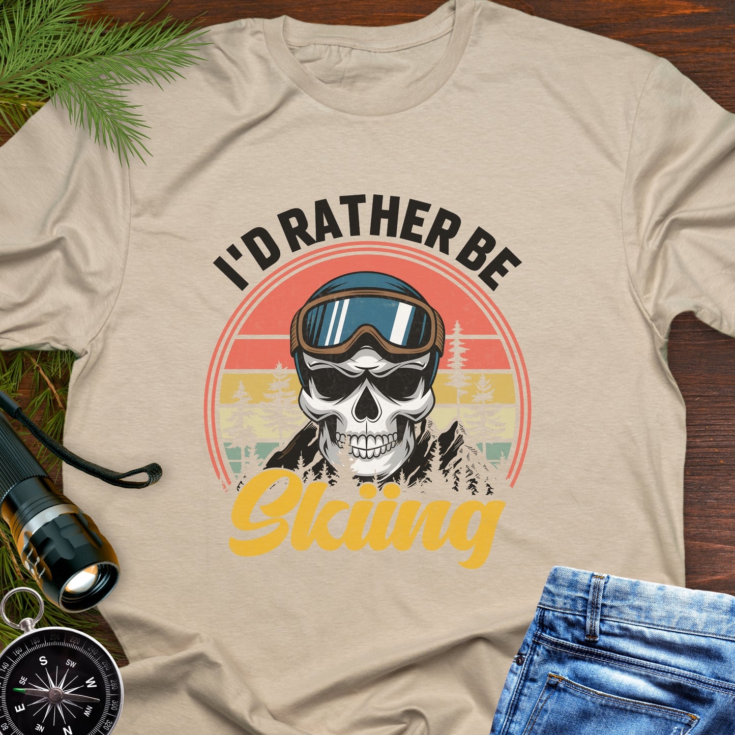 Beige T-shirt graphic showing a skeleton head wearing ski goggles in front of a retro sunset with mountains and trees, with text that reads “I’d Rather Be Skiing.