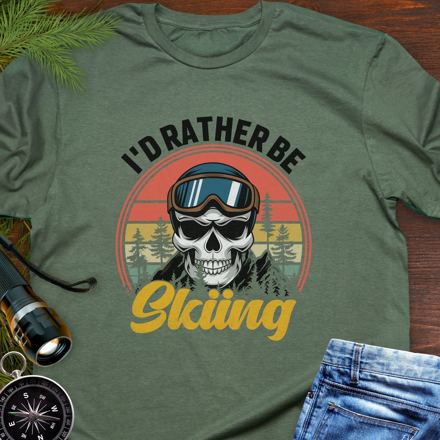 Green T-shirt graphic showing a skeleton head wearing ski goggles in front of a retro sunset with mountains and trees, with text that reads “I’d Rather Be Skiing.