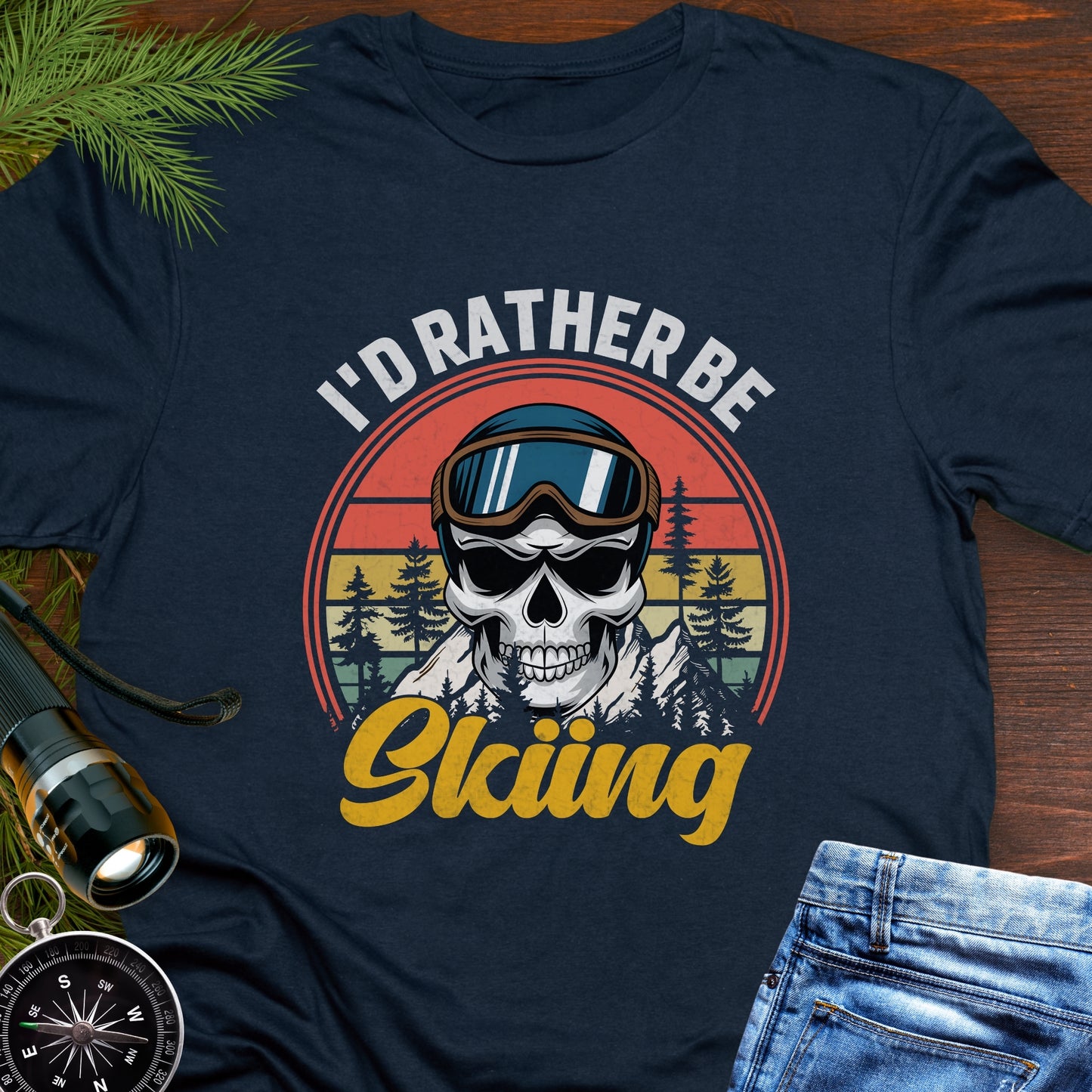 Navy blue T-shirt graphic showing a skeleton head wearing ski goggles in front of a retro sunset with mountains and trees, with text that reads “I’d Rather Be Skiing.