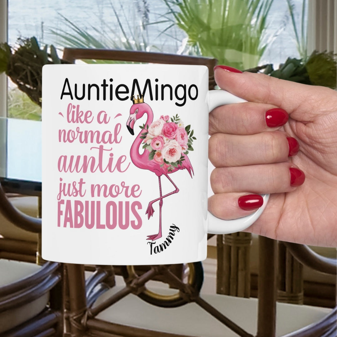 Mug with flamingo design and text held by a hand with red nails, outdoor setting.