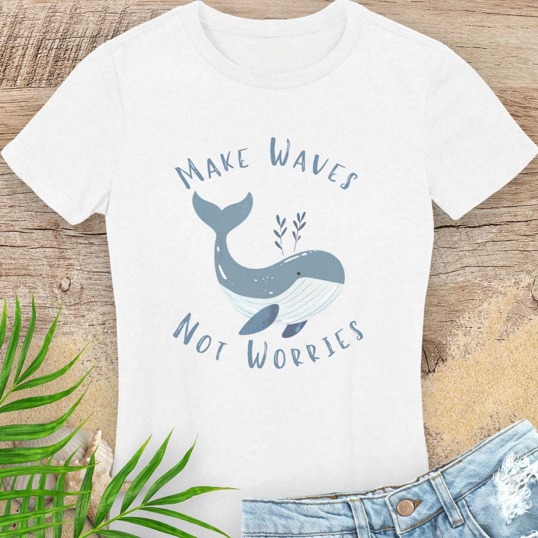 White t-shirt with whale graphic and 'Make Waves Not Worries' text on a wooden surface with leaves and jeans.