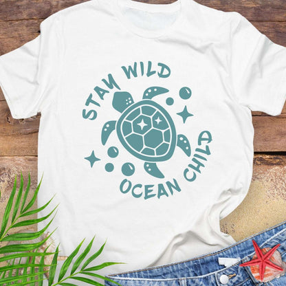 White t-shirt with teal sea turtle graphic and 'Stay Wild Ocean Child' text on a wooden surface with leaves and starfish.