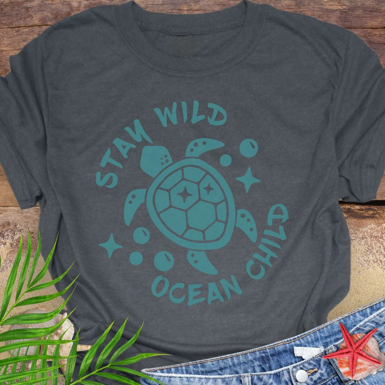 Gray t-shirt with teal sea turtle graphic and 'Stay Wild Ocean Child' text on a wooden surface with leaves and starfish.