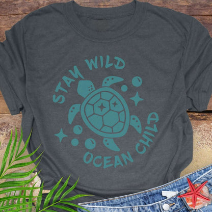 Gray t-shirt with teal sea turtle graphic and 'Stay Wild Ocean Child' text on a wooden surface with leaves and starfish.