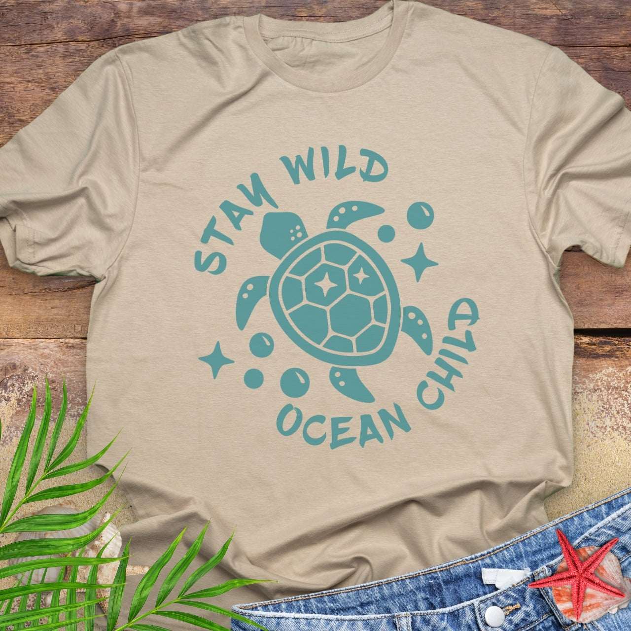 Beige t-shirt with teal turtle graphic and 'Stay Wild Ocean Child' text on a wooden surface.