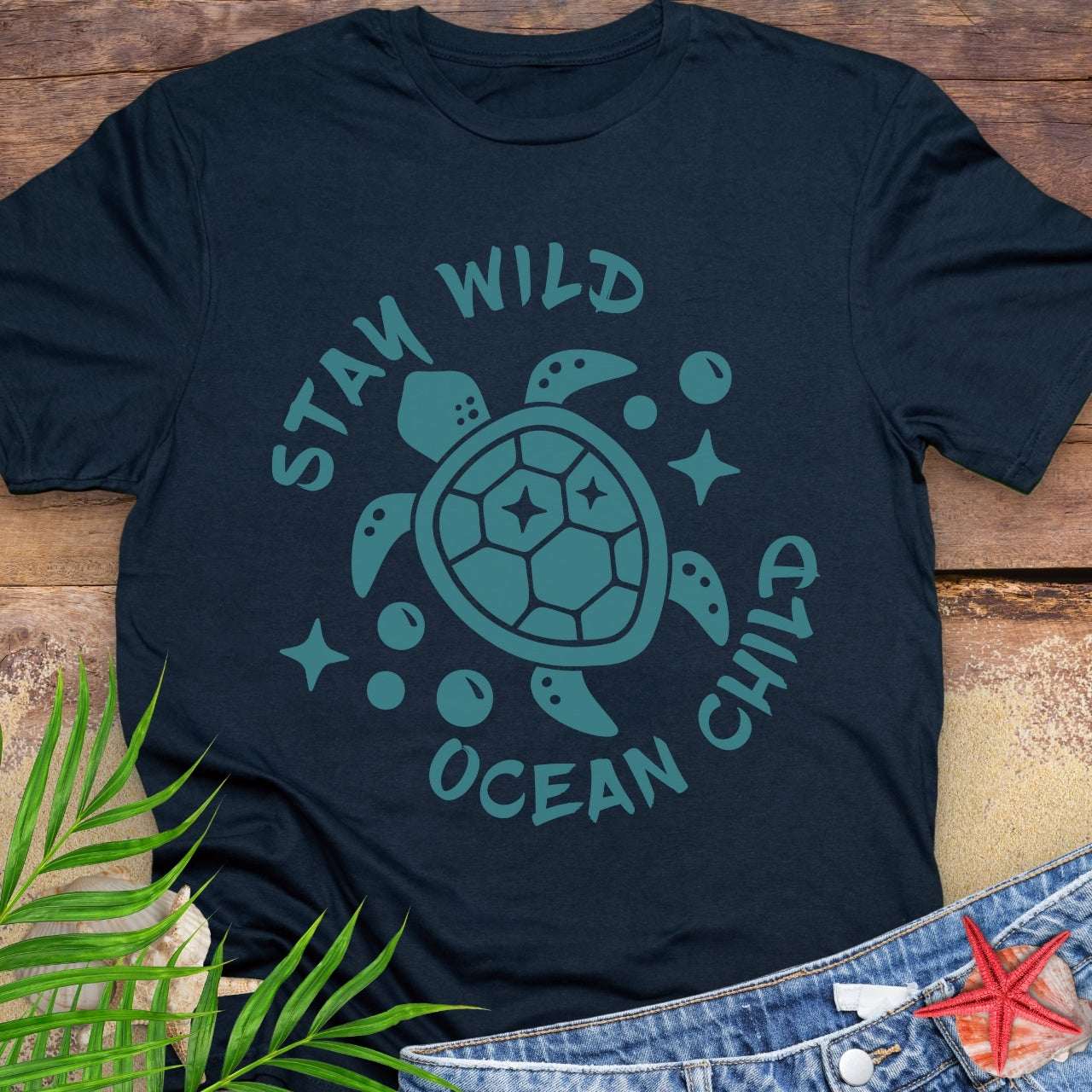 Navy blue t-shirt with teal sea turtle graphic and 'Stay Wild Ocean Child' text on a wooden surface.