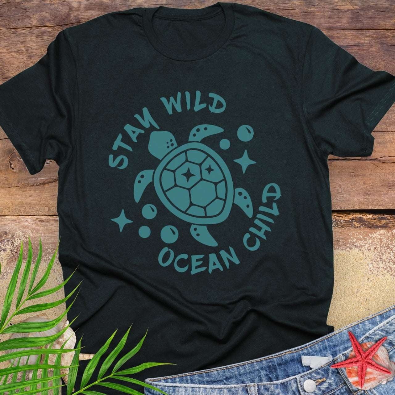 Black t-shirt with teal sea turtle graphic and 'Stay Wild Ocean Child' text on a wooden surface with leaves and jeans.